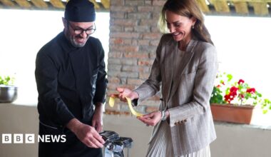 An Italian chef dressed in all black kitchen wear and hat stands to the left of the image next to the Princess of Wales as he helps her make fresh pasta in a kitchen. The Royal, dressed in a grey blazer and skirt is grinning as she holds the pasta in her hands