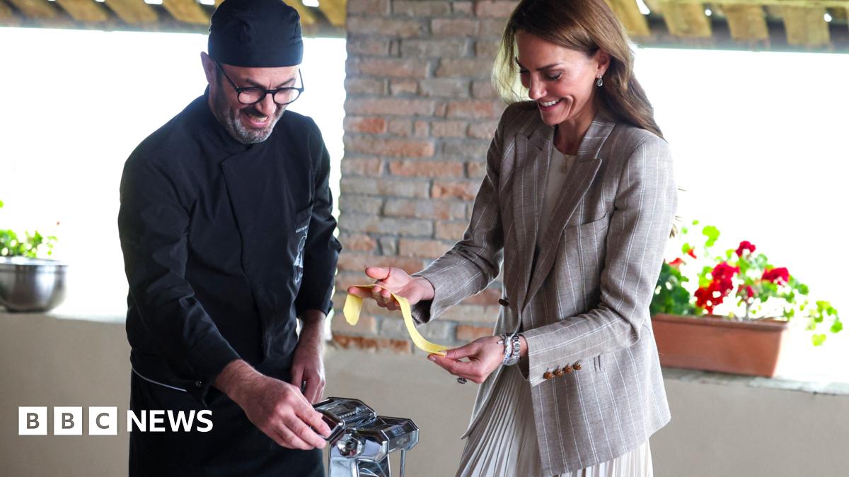 An Italian chef dressed in all black kitchen wear and hat stands to the left of the image next to the Princess of Wales as he helps her make fresh pasta in a kitchen. The Royal, dressed in a grey blazer and skirt is grinning as she holds the pasta in her hands