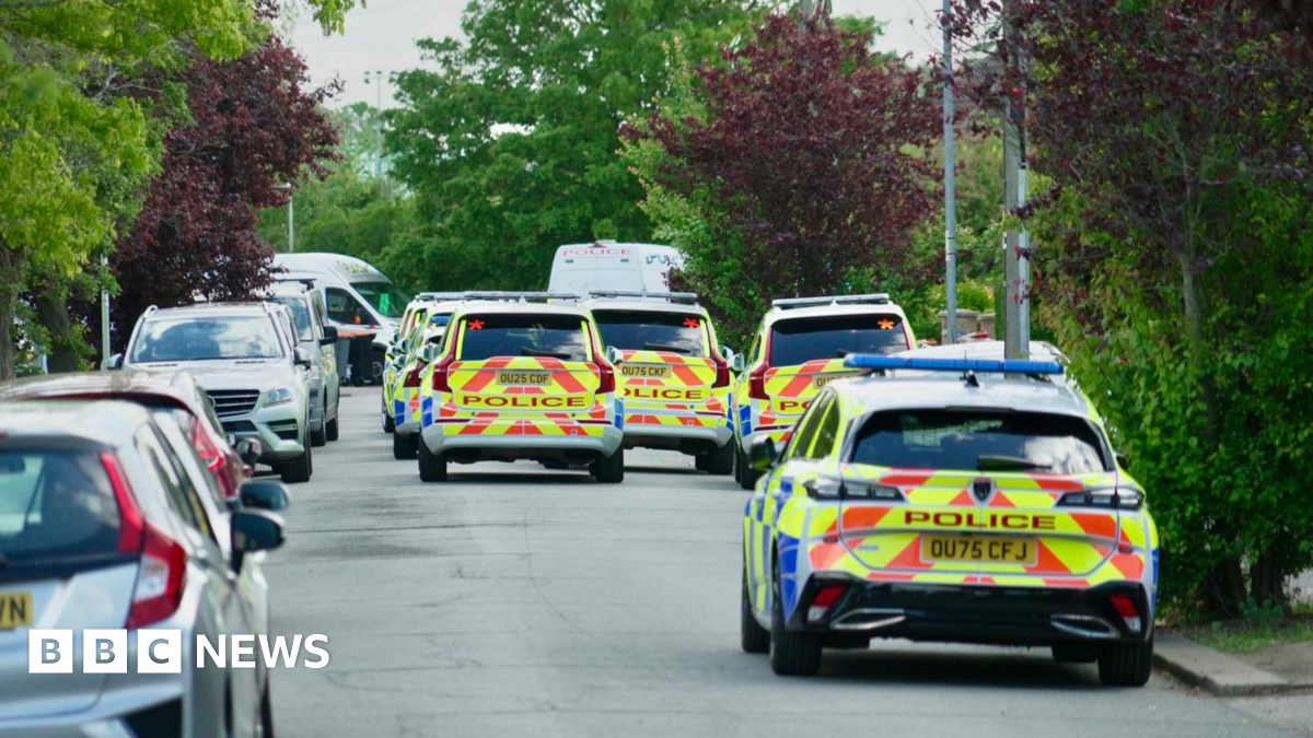 Police cars line a leafy residential street in Bedford - there are other parked vehicles on the left
