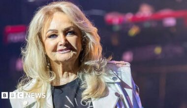 Bonnie Tyler on stage, she has long blonde hair, and is wearing a black T-shirt and a white biker jacket