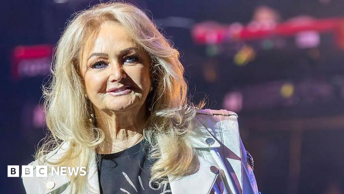 Bonnie Tyler on stage, she has long blonde hair, and is wearing a black T-shirt and a white biker jacket