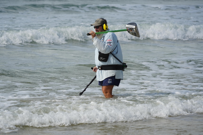 A man carrying a metal detector standing in surf.