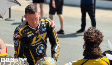 Channing Tatum is wearing black and gold leathers and is leaning over slightly, reaching for a matching helmet, with people stood around him, on a sunny day.