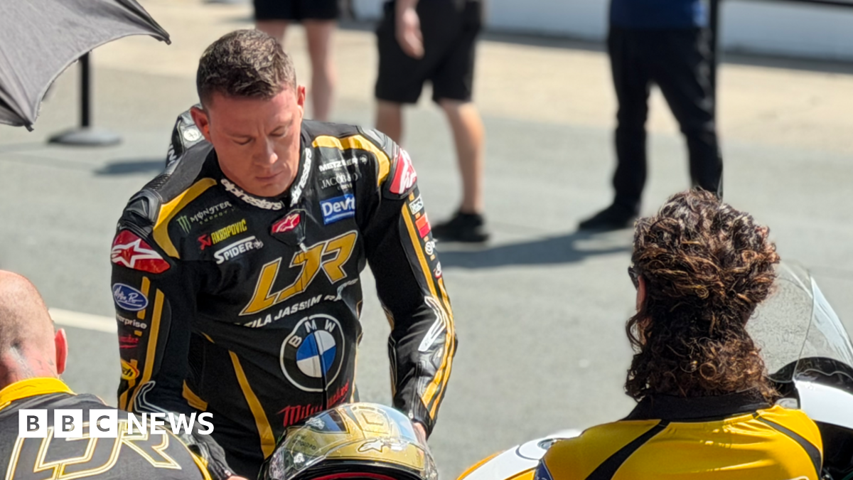 Channing Tatum is wearing black and gold leathers and is leaning over slightly, reaching for a matching helmet, with people stood around him, on a sunny day.
