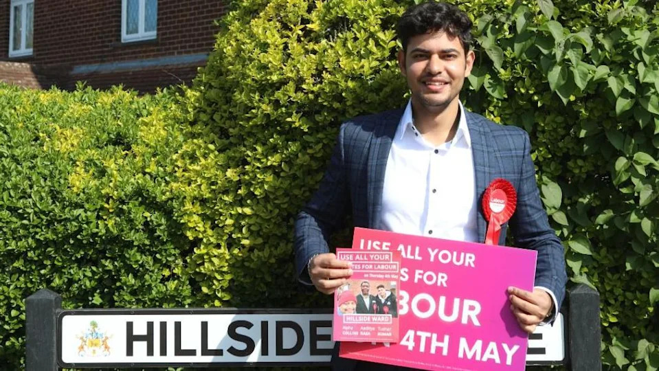 A neatly dressed person, Tushar Kumar, stands on a quiet residential street, framed by lush green hedges. The sky above is bright and clear blue, suggesting a calm, sunny day. Behind Kumar, a street sign reads “HILLSIDE AVENUE,”
Kumar is holding a bold pink campaign sign with white lettering that reads “USE ALL YOUR VOTES FOR LABOUR 4TH MAY,” along with a leaflet featuring smaller images and additional names and text.