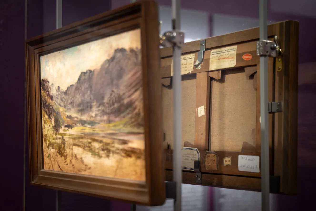 Two paintings facing each other, one of a landscape and one showing the back of a canvas with labels.