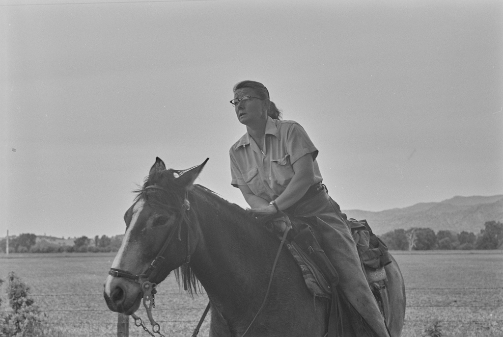 A woman wearing glasses rides a horse across a flat, open valley with mountains and trees visible in the distance.