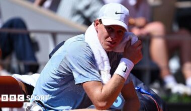 Jannik Sinner, wearing a grey t-shirt and white cap, sits in his chair with a towel around his neck wearing a pained expression
