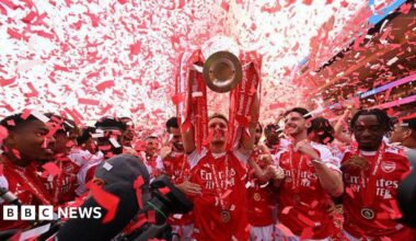 An Arsenal player lifts the Premier League trophy above his head as red and white confetti falls around him, with teammates in red shirts celebrating alongside.