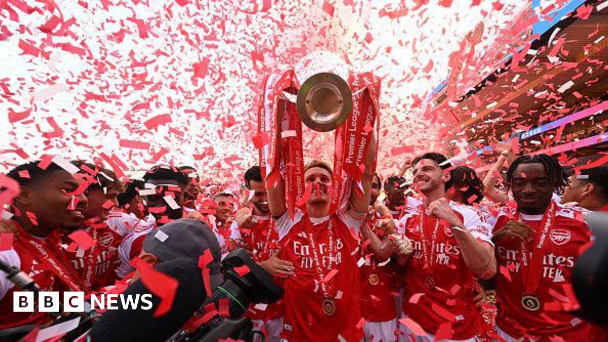 An Arsenal player lifts the Premier League trophy above his head as red and white confetti falls around him, with teammates in red shirts celebrating alongside.
