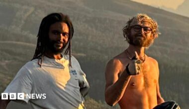 Two young men stand in front of a grassy hilly area and smile at the camera. One wears a white T-shirt and has black dreadlocks. He holds his bike in one hand while resting his other arm behind his back. The other has curly blonde hair, is shirtless, has glasses on and raises his thumb. He sits on top of his bike.