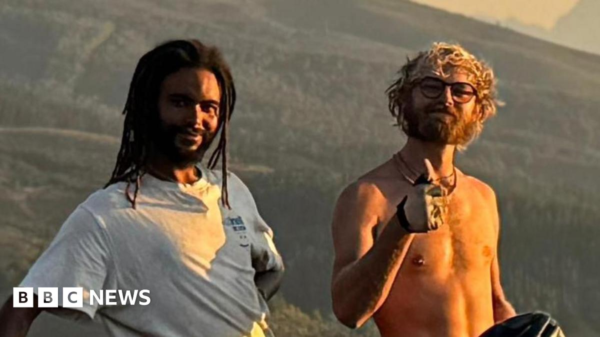 Two young men stand in front of a grassy hilly area and smile at the camera. One wears a white T-shirt and has black dreadlocks. He holds his bike in one hand while resting his other arm behind his back. The other has curly blonde hair, is shirtless, has glasses on and raises his thumb. He sits on top of his bike.