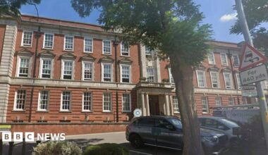 A picture of the three-storey brick council house building on a sunny day.  Pollarded trees are in front of the car park.