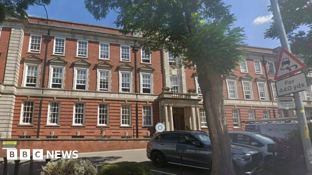 A picture of the three-storey brick council house building on a sunny day.  Pollarded trees are in front of the car park.