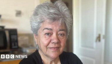 Liz is looking at the camera. She has short grey hair. She is wearing a black cardigan and blue patterned top.