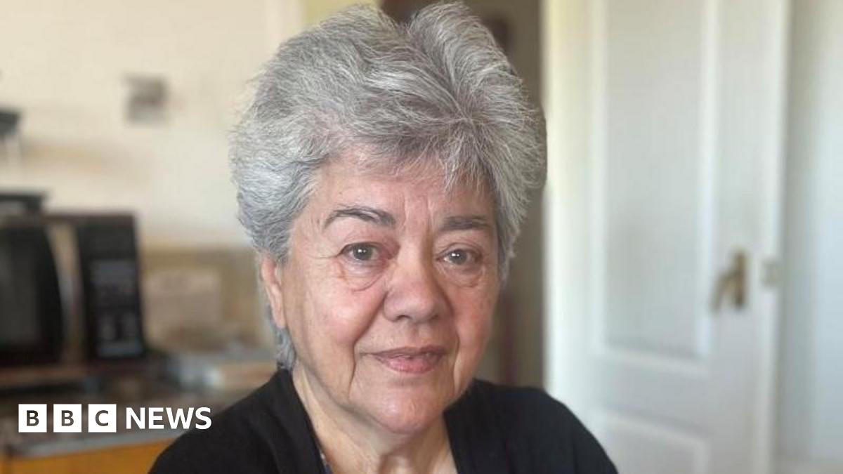 Liz is looking at the camera. She has short grey hair. She is wearing a black cardigan and blue patterned top.