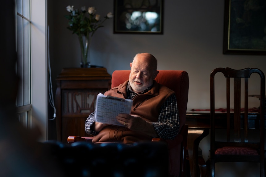 An older man writing on a clipboard as he sits in a lounge room.