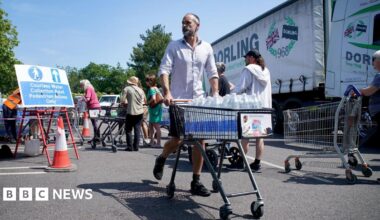A man in a white shirt and shorts is pushing a trolley full of water bottles. People are queuing behind him and a lorry is in the background.