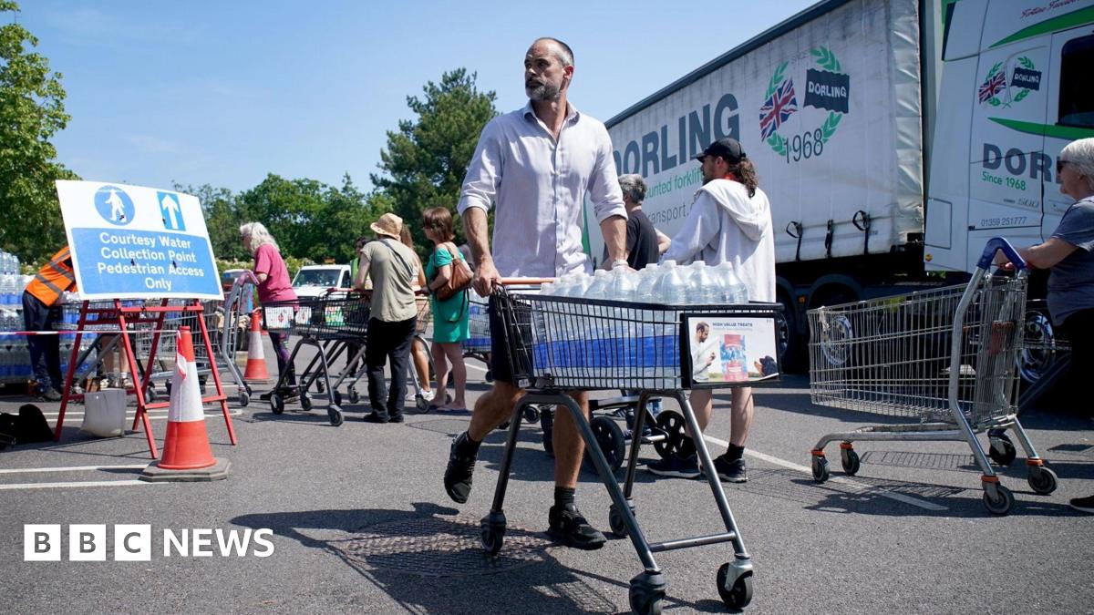 A man in a white shirt and shorts is pushing a trolley full of water bottles. People are queuing behind him and a lorry is in the background.