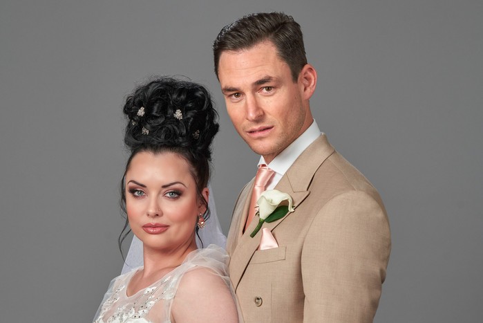 Will Whitney and Zack get married? Whitney and Zack are posed facing the left, but looking into camera. Whitney (left) is in a wedding dress. Her hair is tied up decoratively above her head. Zack (right) is wearing a light beige suit. They're holding hands.