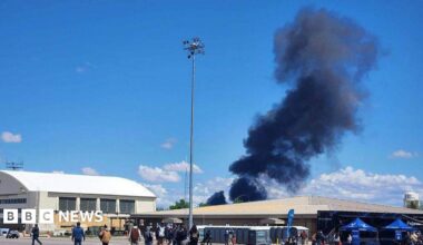 Black smoke rises into a blue sky over two buildings at a US military base.