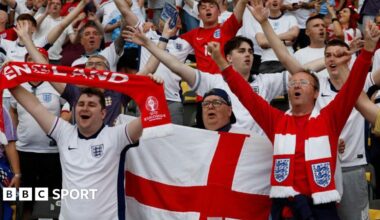 England fans before the Euro 2024 final, many wearing white team shirts, while some have scarves and one holds a red and white Cross of St George flag