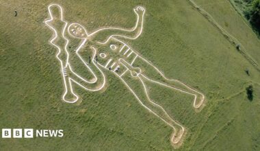An aerial shot of a white outline of a club-wielding naked man on a green grass hill. People are seen dotted around the white figure, some are crouching on the ground and others are walking and carrying some kits in their hands.