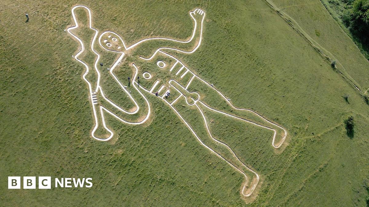 An aerial shot of a white outline of a club-wielding naked man on a green grass hill. People are seen dotted around the white figure, some are crouching on the ground and others are walking and carrying some kits in their hands.