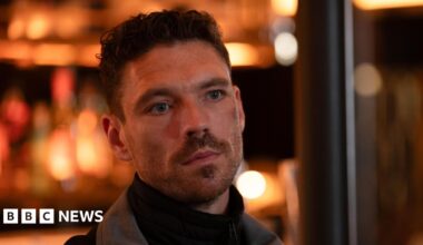 A man in a roll neck black jumper and grey jacket - inside a bar with blurry lights behind. He has facial hair and curly cropped hair.