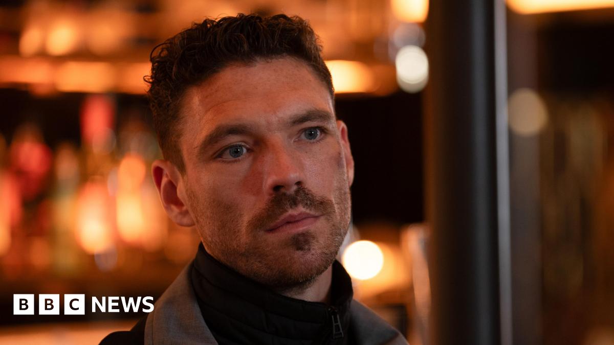 A man in a roll neck black jumper and grey jacket - inside a bar with blurry lights behind. He has facial hair and curly cropped hair.