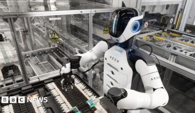The Aeon robot at work in the factory. It has a white torso with black shoulders. Its black plastic face has a vertical line of blue light.