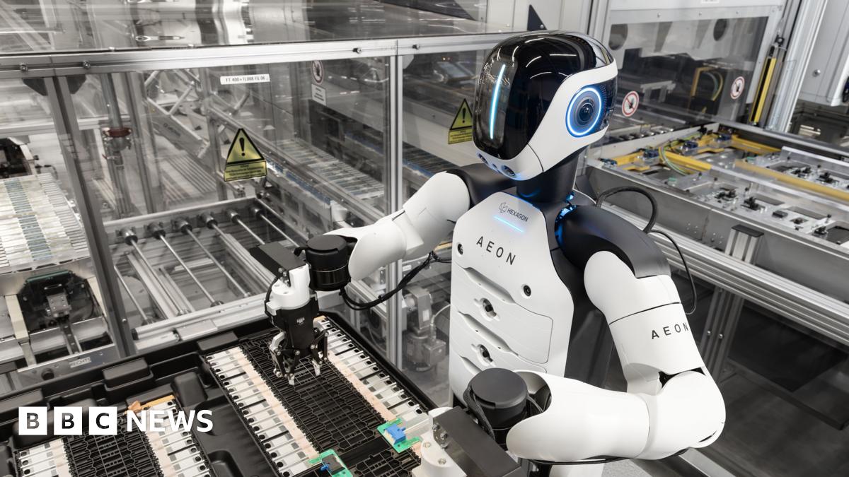 The Aeon robot at work in the factory. It has a white torso with black shoulders. Its black plastic face has a vertical line of blue light.