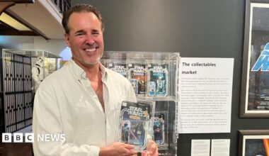 Matt Fox wears a white shirt and is standing in front of a wall of boxed Star Wars figurines. He is holding a Darth Vader box.