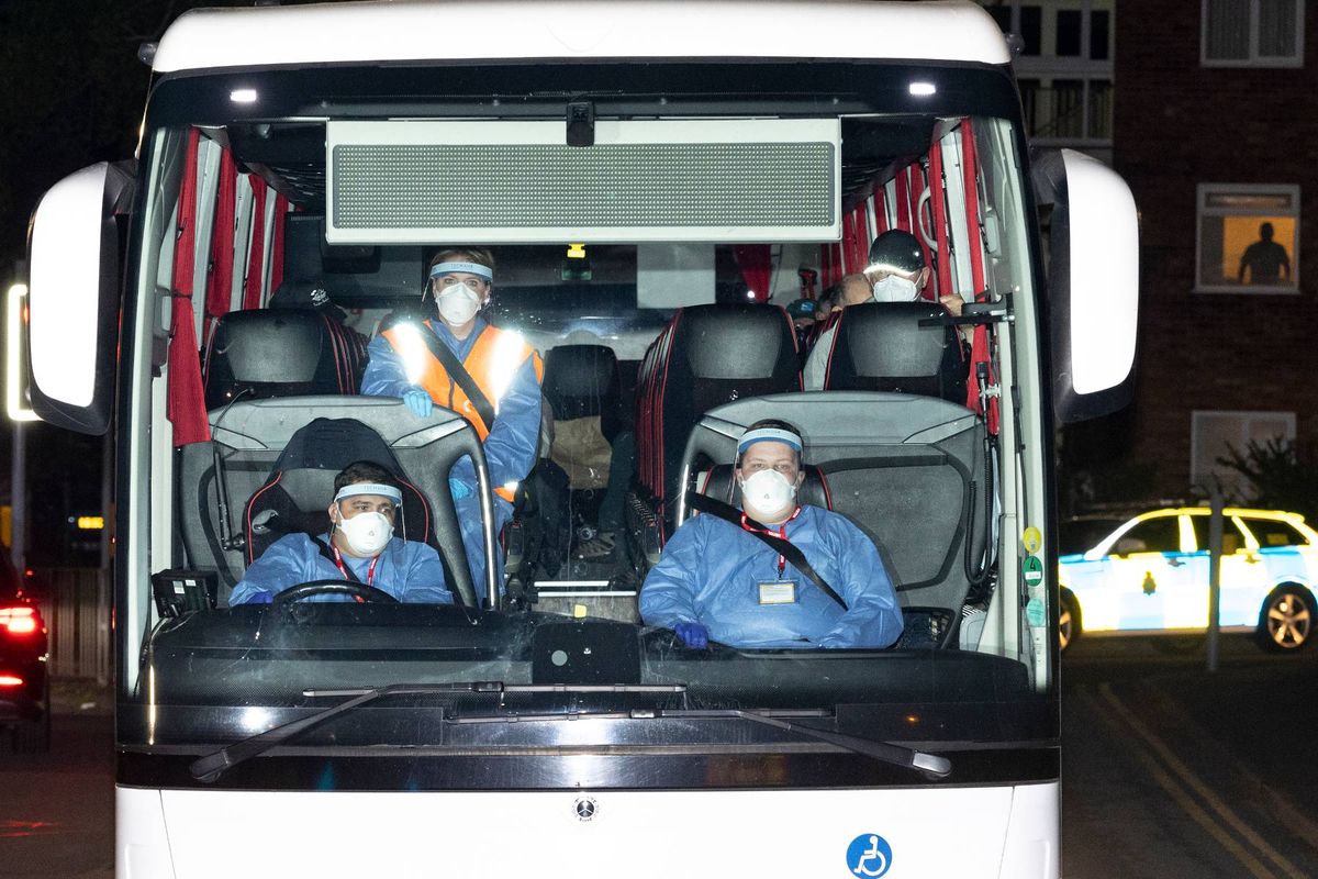 An image depicts the interior of a bus with several individuals seated inside. The occupants are wearing protective gear, including masks and possibly gloves. The bus is parked on the side of a road, with a car visible in the background, indicating a nighttime setting.