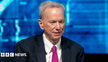 Eric Schmidt sits with his hands crossed and looks ahead. He is wearing a black suit, a white shirt, and a hot pink polka-dot tie. In the background is a blue screen that features different shades of blue.