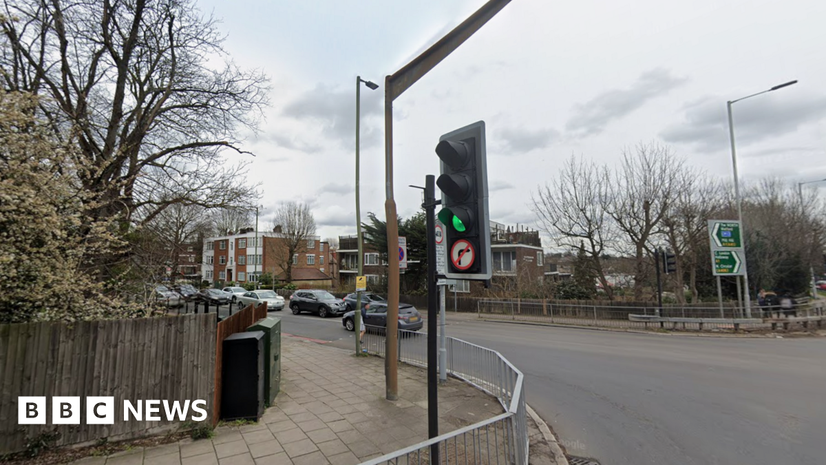 A general view of a pedestrian traffic light crossing on Holders Hill Road, with cars on the road and residential buildings and trees in the background.