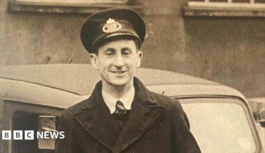 A black and white image of a postman in the post-war era in the late 1940s. He wears a long black coat, black hat, tie, white shirt and is standing in front of a car.