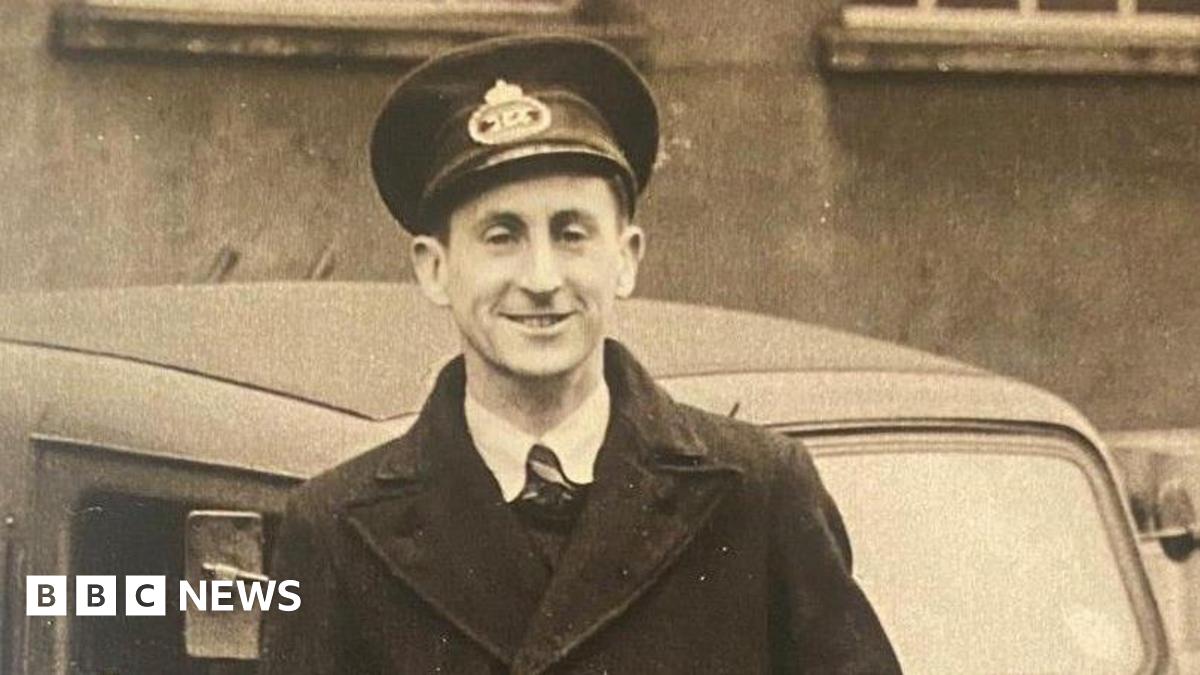 A black and white image of a postman in the post-war era in the late 1940s. He wears a long black coat, black hat, tie, white shirt and is standing in front of a car.