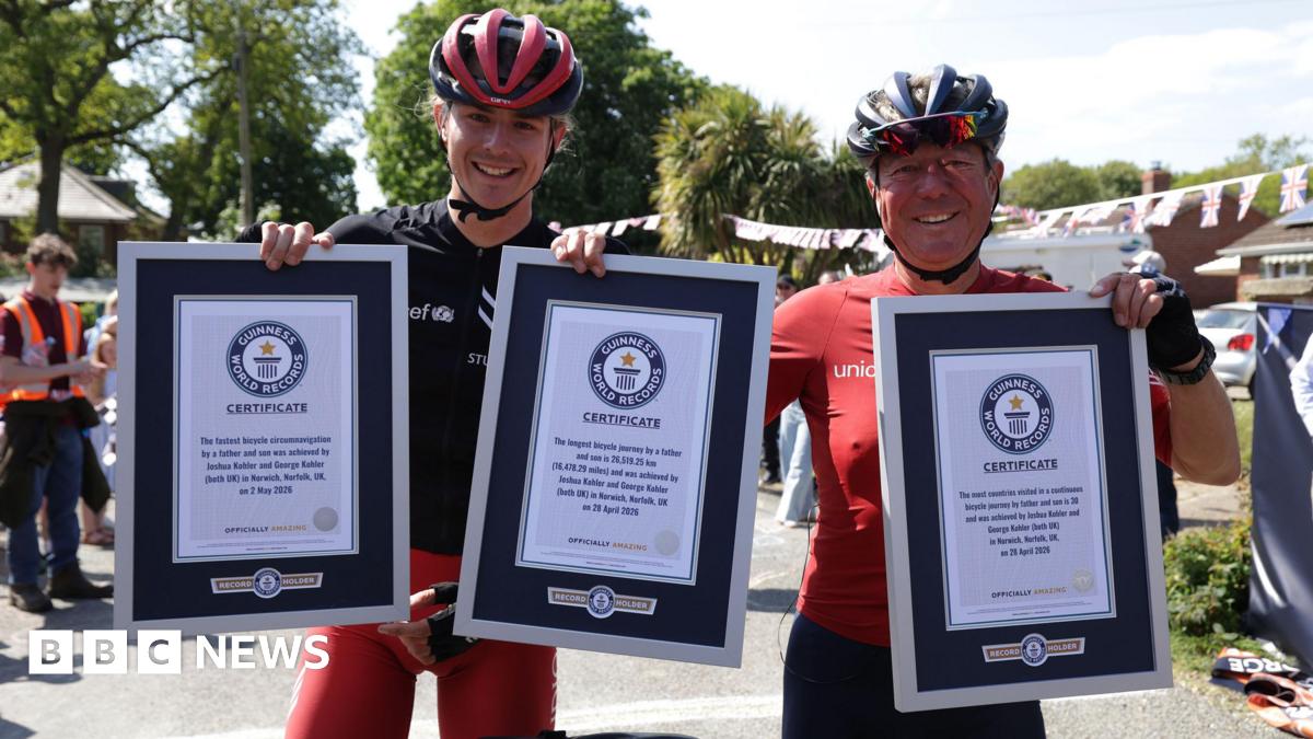 Josh and George Kohler are standing next to each other. They are both wearing red and black cycling wear and helmets. They are holding up three Guinness World Record certificates.