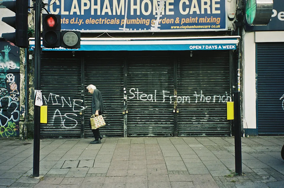 Colour photograph of an elderly person walking along the street past a shuttered shop front that has “STEAL FROM THE RICH” spray-painted in front