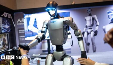 A humanoid robot being demonstrated at an event. With the stance of a human, its arms, torso and pelvis are silver brushed metal, with black joints and black hands. The face is a hollow ring that glows icy blue around the perimeter. In the background is a display board with large images of the same robot, while human hands reach out to shake the robot's black hands.