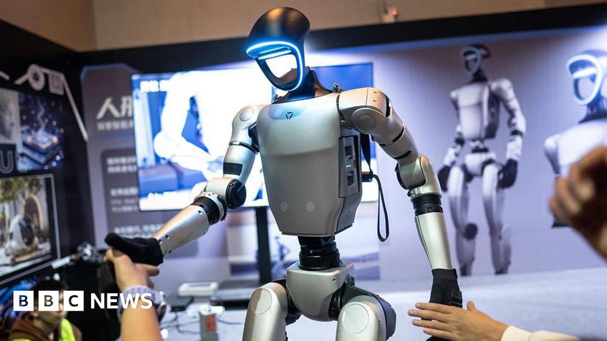 A humanoid robot being demonstrated at an event. With the stance of a human, its arms, torso and pelvis are silver brushed metal, with black joints and black hands. The face is a hollow ring that glows icy blue around the perimeter. In the background is a display board with large images of the same robot, while human hands reach out to shake the robot's black hands.