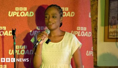 A woman talks into a microphone on stage. She stands in front of a red backdrop which says "BBC Upload" on it.