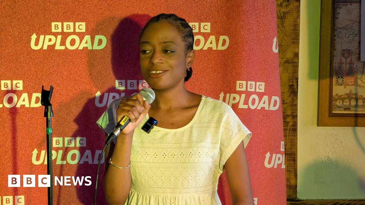 A woman talks into a microphone on stage. She stands in front of a red backdrop which says "BBC Upload" on it.