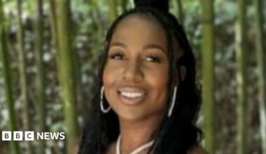 A young woman wearing gold jewellery smiles. She is standing in front of a forest.
