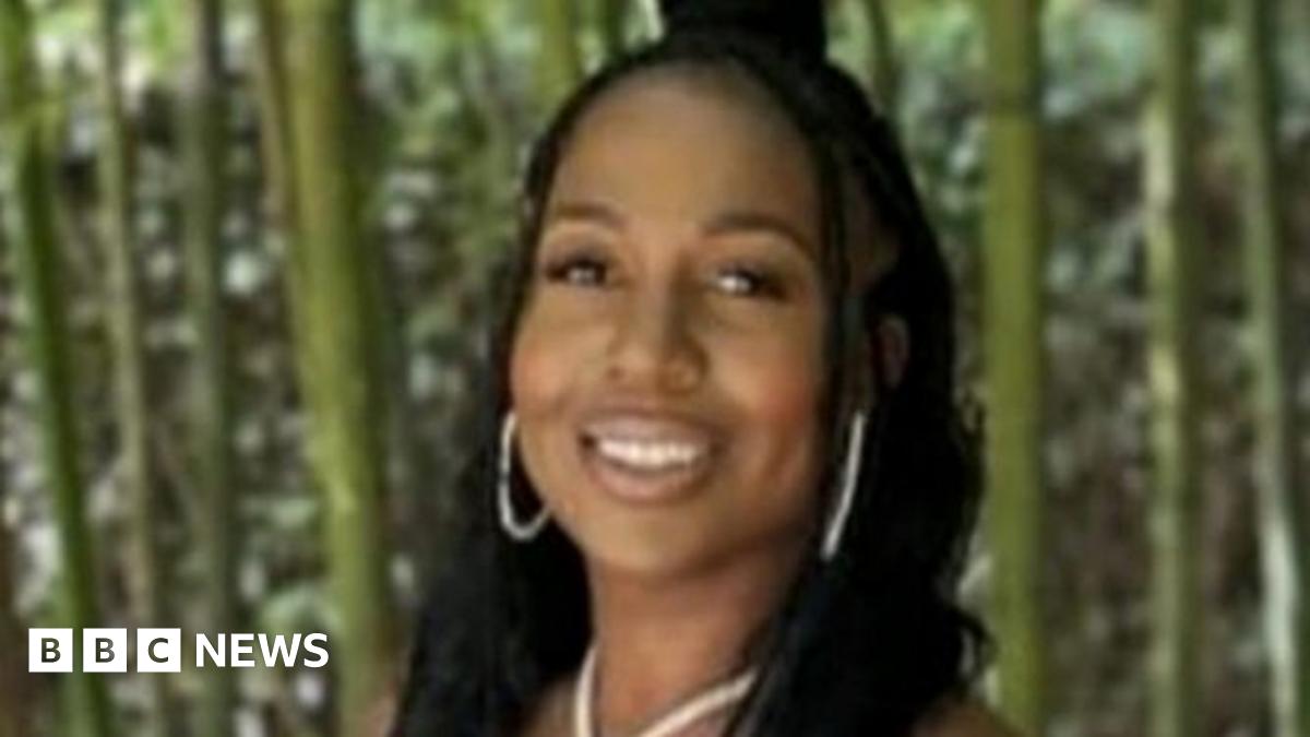 A young woman wearing gold jewellery smiles. She is standing in front of a forest.