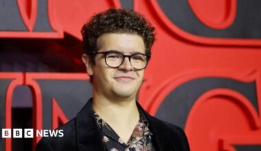 Gaten Matarazzo has short curly hair and is wearing black-rimmed glasses, a black Velvet blazer and floral shirt underneath. He is standing on a red carpet in front of a huge 'Stranger Things' sign
