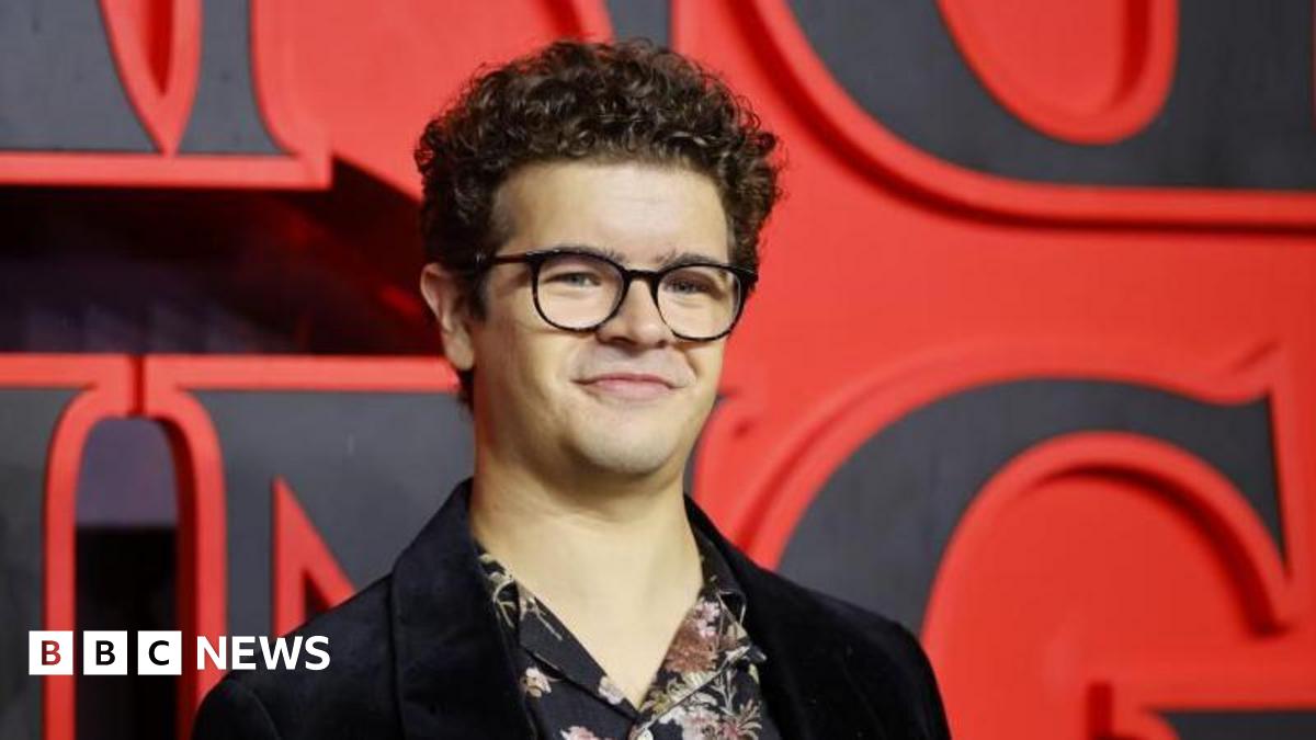 Gaten Matarazzo has short curly hair and is wearing black-rimmed glasses, a black Velvet blazer and floral shirt underneath. He is standing on a red carpet in front of a huge 'Stranger Things' sign