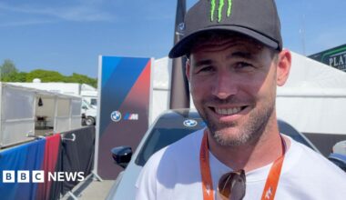 Sir Mark smiling and wearing a black Monster Energy cap, white t-shirt with sunglasses hooked onto the neckline,  and an orange TT lanyard. There are tents and barriers behind him on a sunny day.
