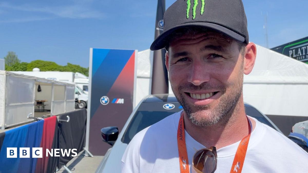 Sir Mark smiling and wearing a black Monster Energy cap, white t-shirt with sunglasses hooked onto the neckline,  and an orange TT lanyard. There are tents and barriers behind him on a sunny day.